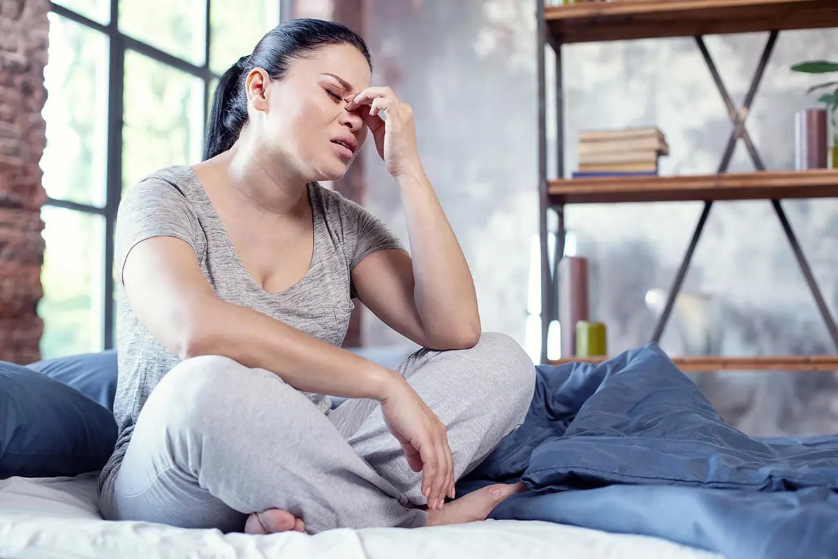 Una mujer de edad media sentada en la cama, se toca la cabeza en señal de dolor y necesita opciones de cómo evitar el insomnio en la menopausia.